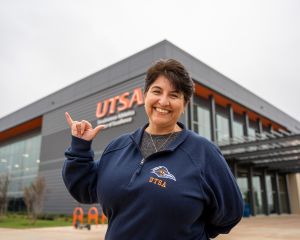 UTSA Online alum Brenda Davila holding up a Roadrunner sign while standing in front of the UTSA Race building
